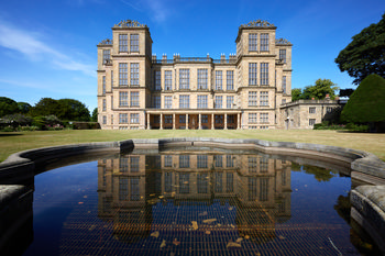 Harwick Hall pond The image is an architectural photograph featuring the Elizabethan manor house of Hardwick Hall, which is located in Derbyshire, United Kingdom. In the foreground, the clear and still pond reflects the grand facade of Hardwick Hall, capturing detailed architectural elements such as its numerous windows and rooftop parapets. The scene is set on a summer morning, as evidenced by the bright sunlight and blue skies. Hardwick Hall is a prominent landmark managed by the National Trust and is renowned for its Elizabethan design. The photograph highlights the relationship between the manor house and the pond, showcasing the symmetry and landscaped gardens typical of estate grounds in the United Kingdom.